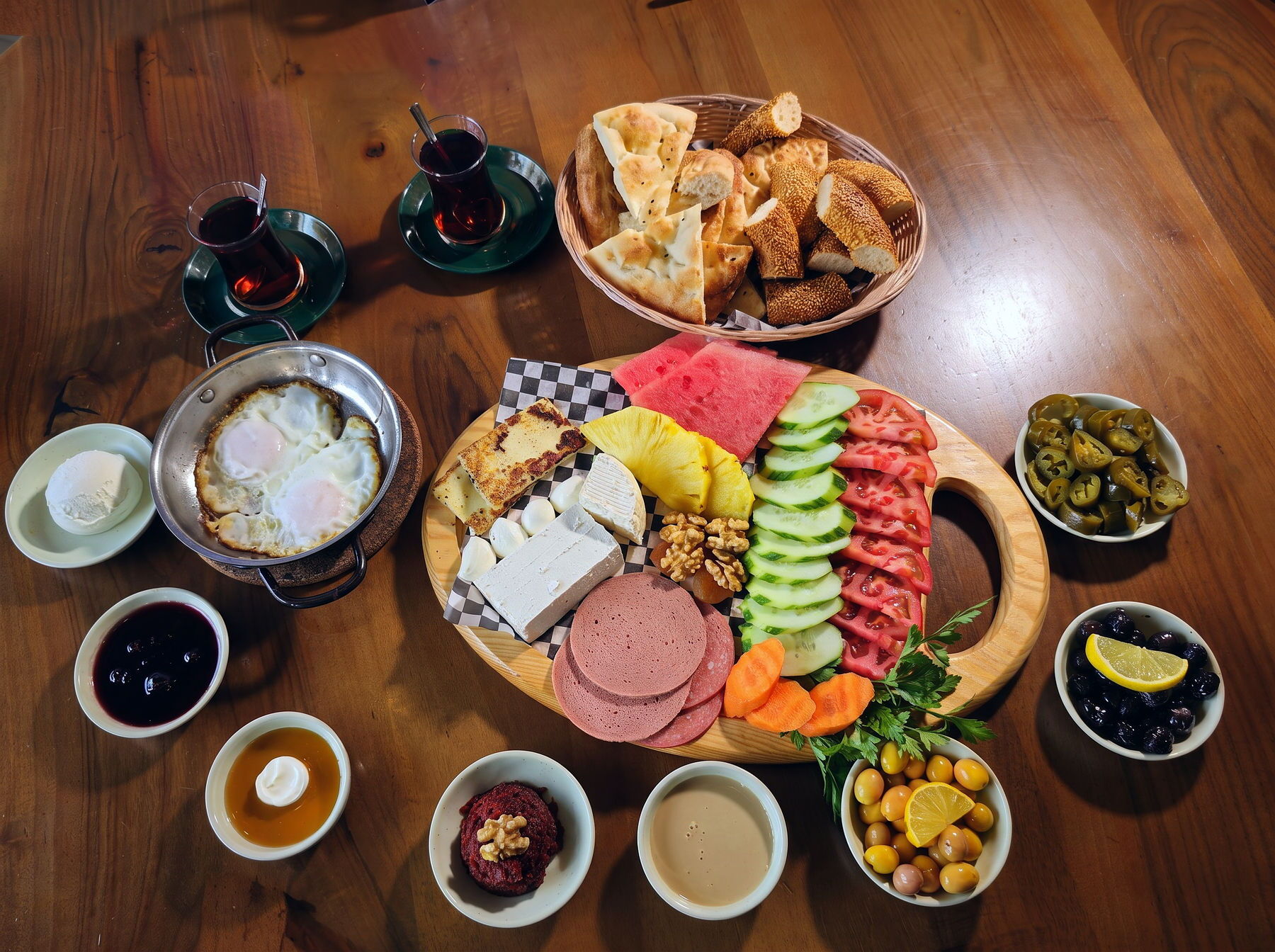 An overhead Turkish breakfast for two on a dark wooden table: a board of cheeses, tomatoes, cucumber, melon, walnuts and cold meats, ringed by bowls of olives, honey, pekmez, clotted cream and pickles, with a pan of fried eggs, simit and two glasses of cay.