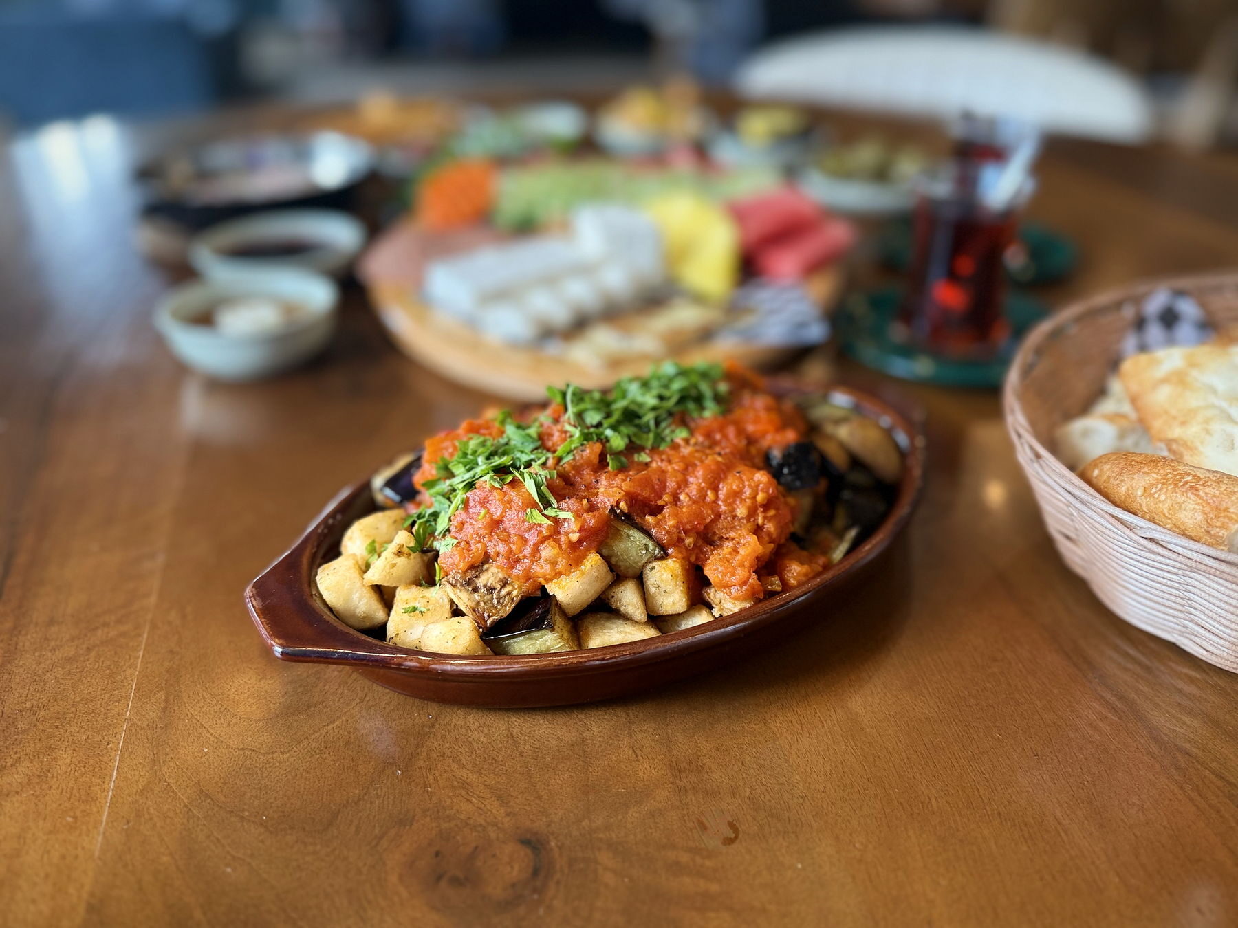 An oval terracotta dish of crispy fried eggplant and potato under a red house sauce, topped with chopped parsley, on a wooden table beside a basket of bread, with the rest of a breakfast service blurred behind.