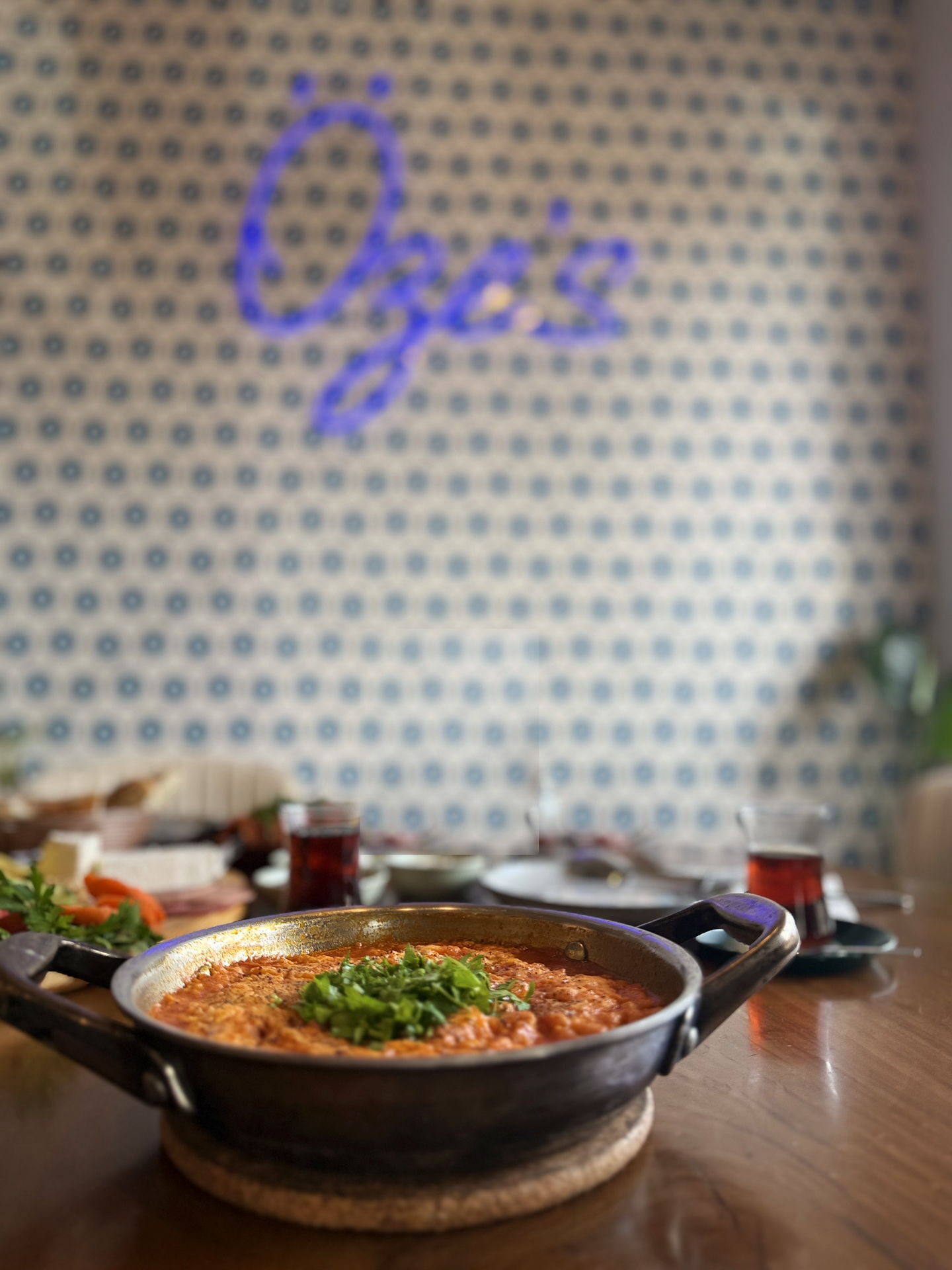 A steel pan of menemen topped with chopped parsley on the café counter, out of focus behind it, the Öze's neon sign glowing blue over a wall of patterned tile.