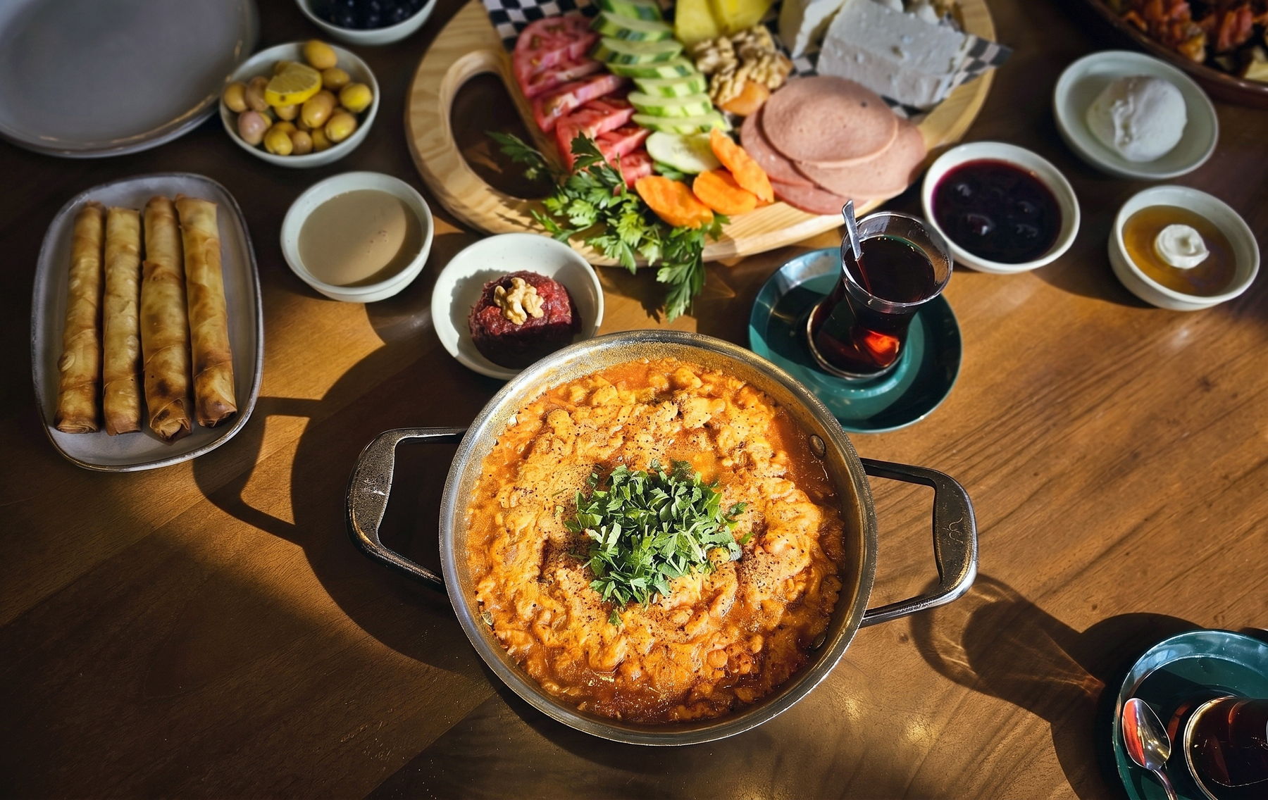 A steel pan of menemen crowned with chopped parsley on a sunlit wooden table, ringed by the full Turkish breakfast: a board of cheeses and cold cuts, sigara borek, olives, preserves and two glasses of cay, photographed from above.