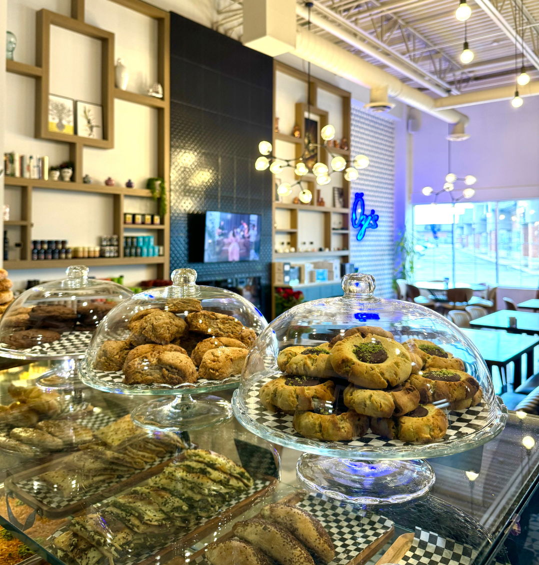 The cafe's glass pastry counter, cookies and pistachio-topped rings under domed cloches, with the shelved back wall, pendant lights and the blue Öze's neon sign behind.