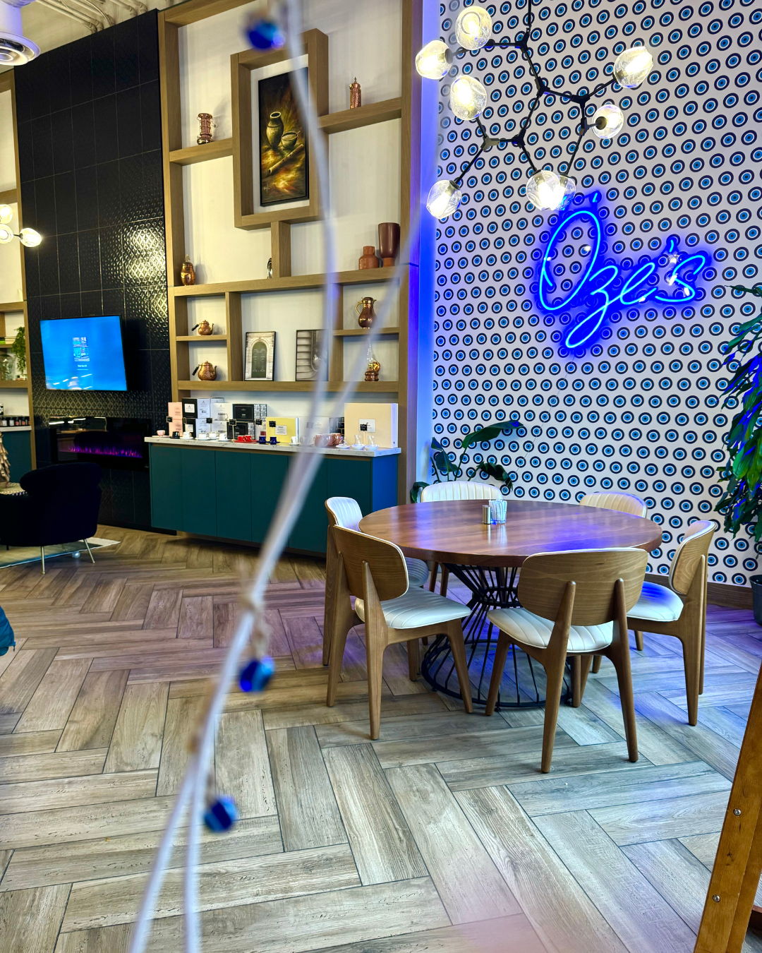 A round wooden table and four pale chairs on a herringbone floor in front of a wall of blue evil-eye tiles carrying the Öze's neon sign, strung bulbs overhead.