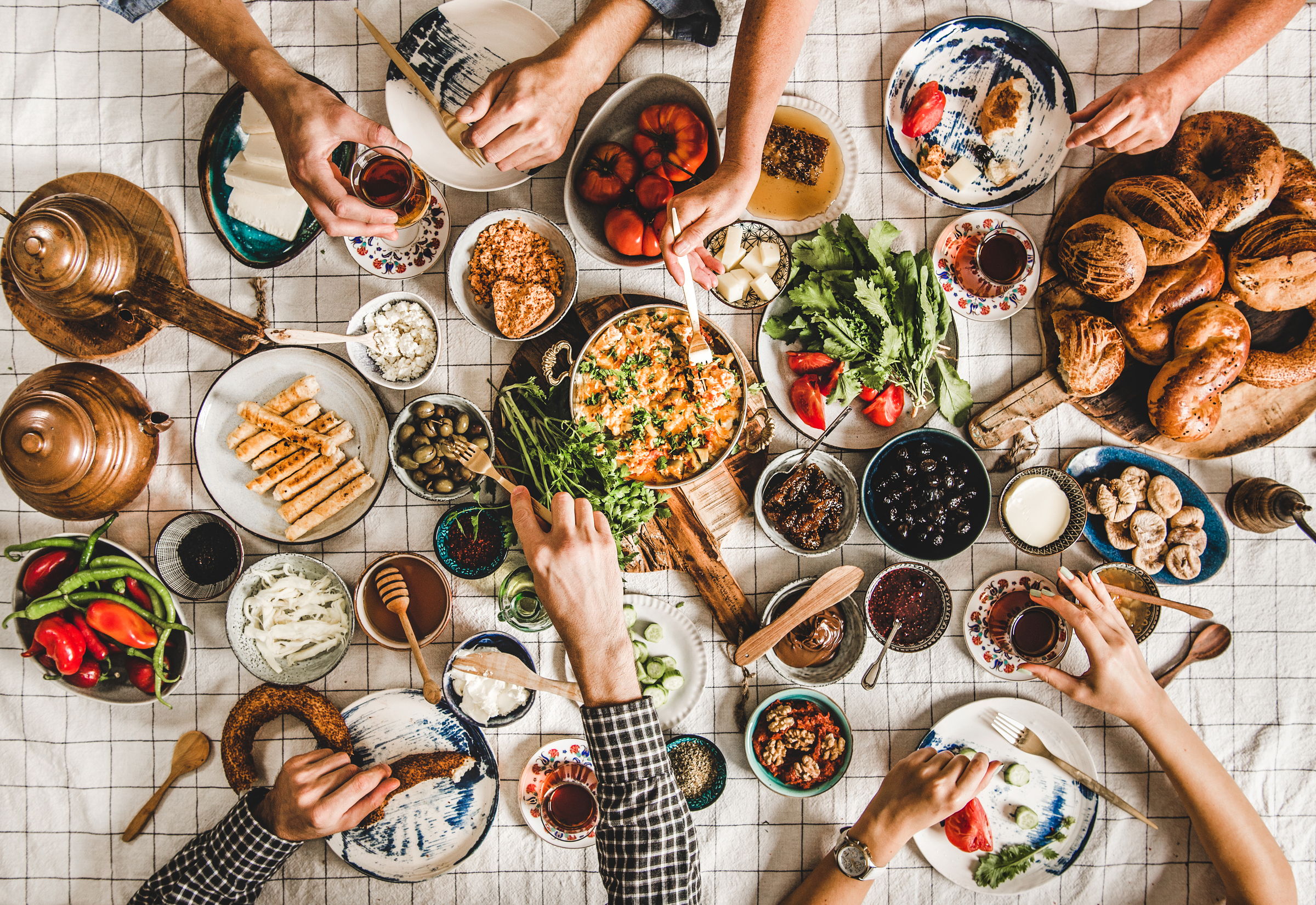 Many hands reaching at once across a crowded breakfast table on a checked cloth — pouring tea from copper pots, tearing simit, passing plates of cheese, olives, tomatoes and eggs.