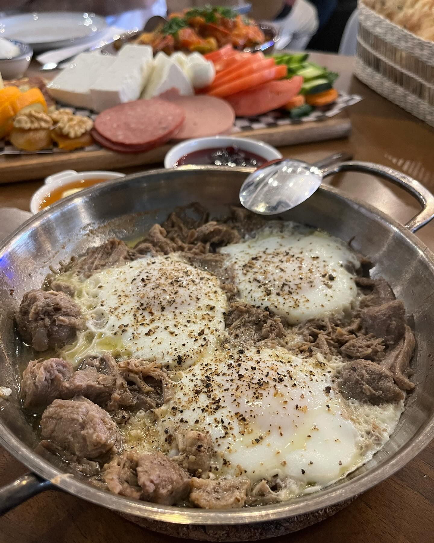 A steel pan of diced meat cooked around three peppered eggs, on a wooden table, with a board of white cheese, sliced sucuk, tomato and cucumber behind it.
