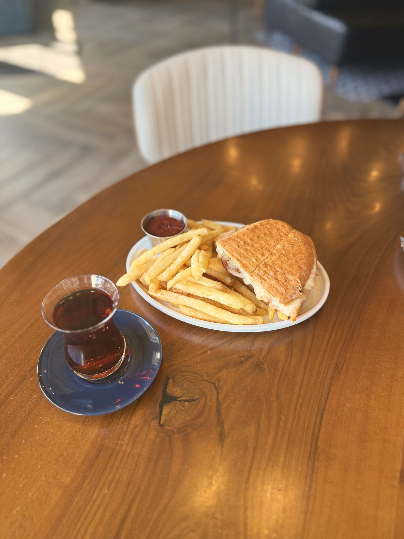 A glass of Turkish tea on a navy saucer beside a plate of pressed toast and golden fries, on a warm wooden table.